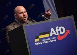 یک مرد میانسال سفید و کچل با انگشت اشاره می کند و روی تریبونی می ایستد که عبارت «AfD» و نوشته آلمانی روی آن نوشته شده است.